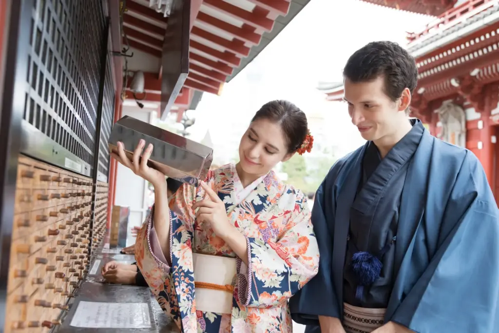 A couple in kimono shaking an omikuji fortune box beside wooden drawers at a red temple hall