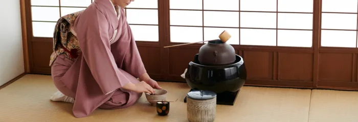 A woman in a pink kimono kneeling beside an iron tea kettle in a tatami room