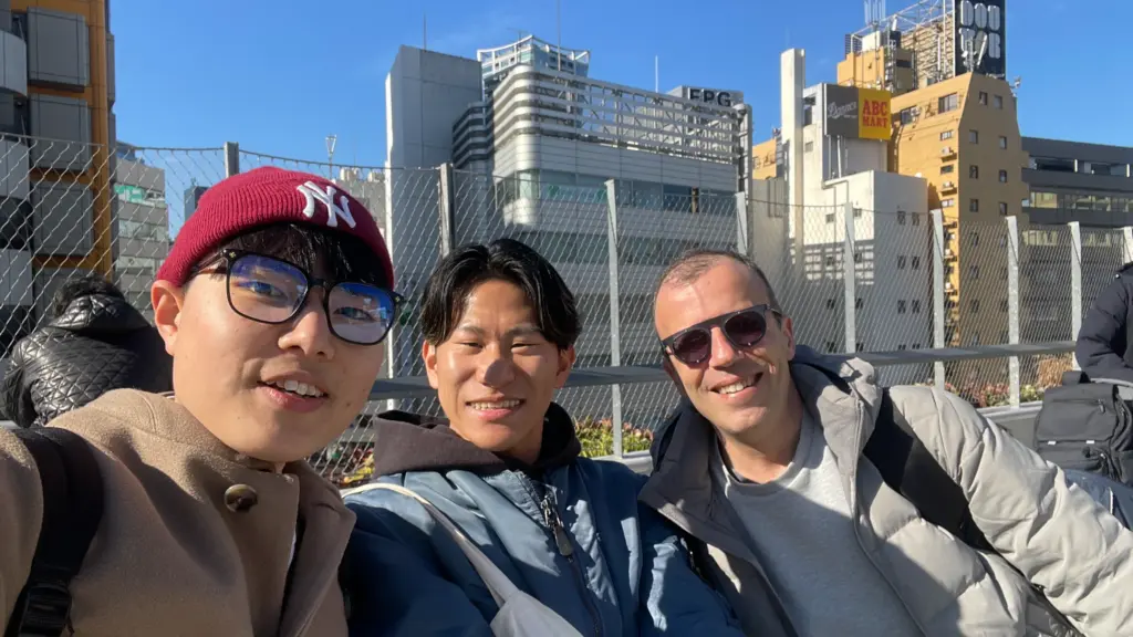 Three people smiling in a selfie on a sunny footbridge above a city street