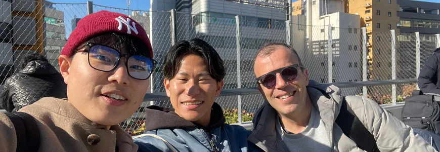 Three people smiling in a selfie on a city footbridge with office buildings behind them