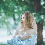 A woman with long blonde hair in a pale dress standing among blurred green trees