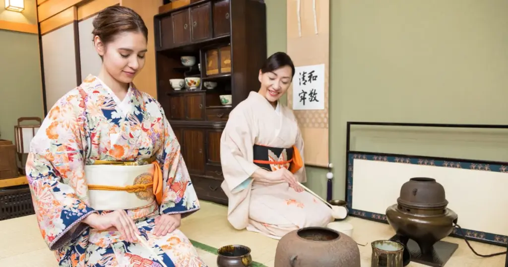 Two women in kimono kneeling in a tatami tea room as one prepares tea beside a hanging scroll